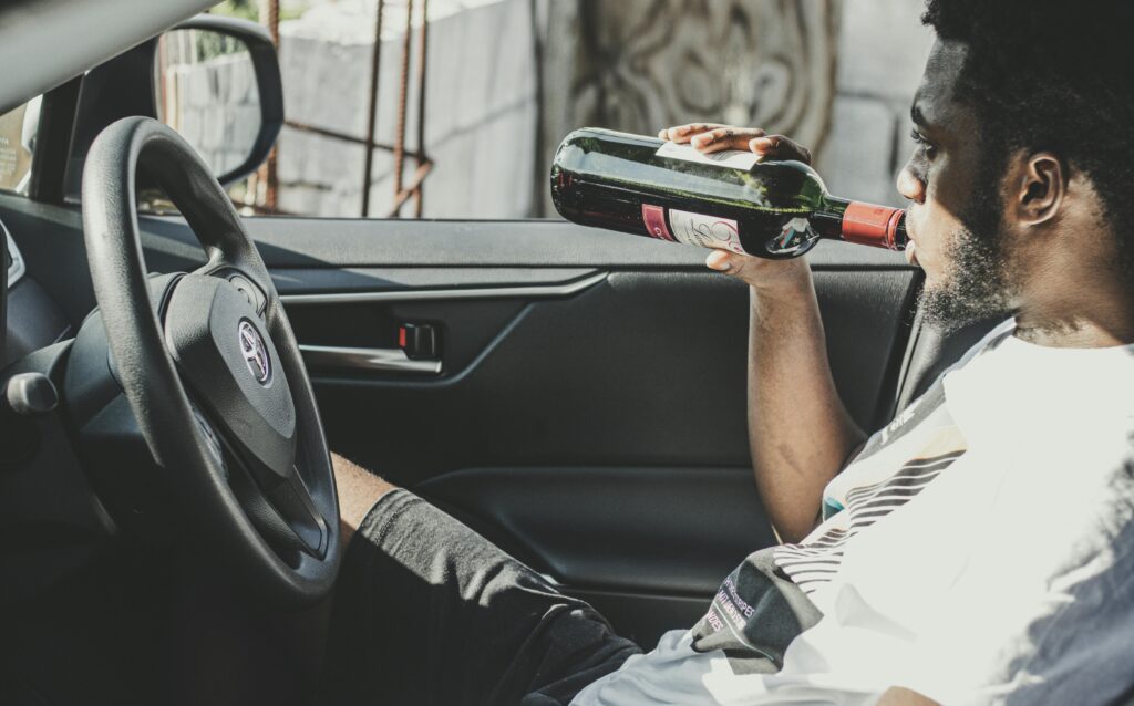 A man sits inside a car drinking red wine from a bottle, side view.