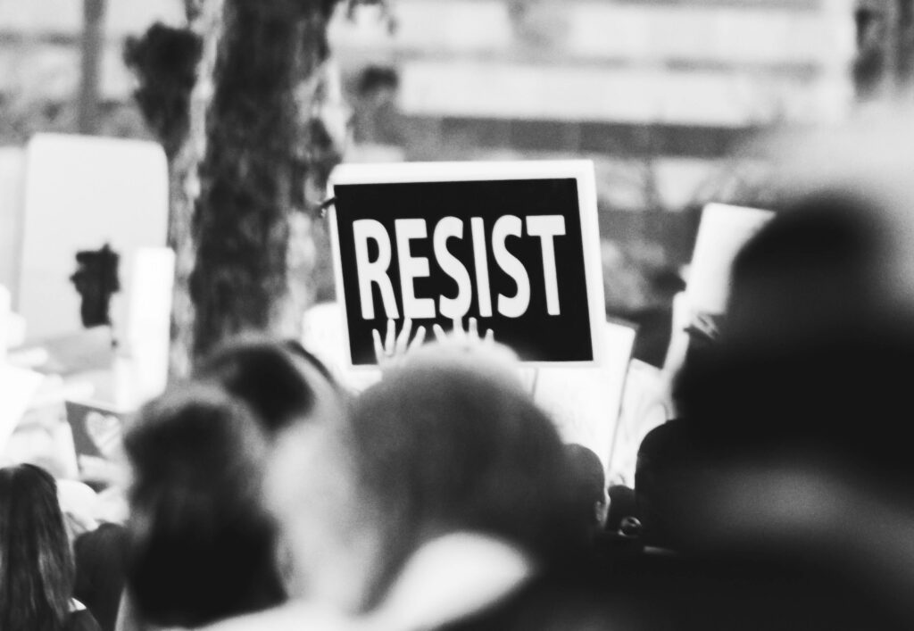 pexels-photo-3141240-3141240 Monochrome image showing a crowd holding a 'Resist' sign at a protest.